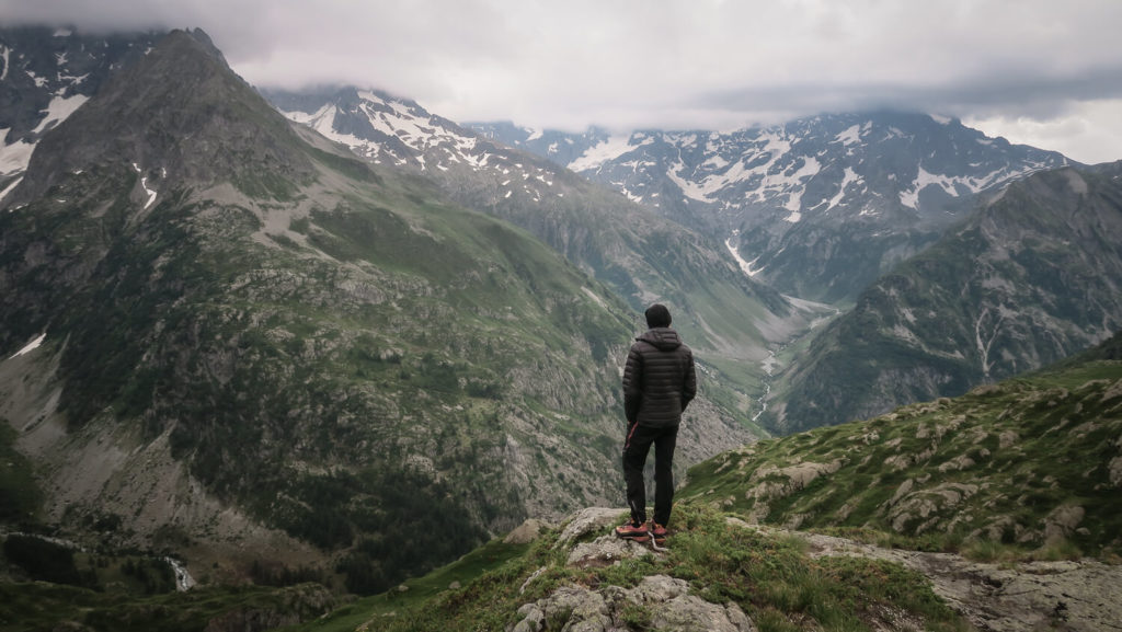 Homme de dos avec doudoune noire, bonnet et chaussures d'alpinisme devant un panorama de montagne (illustration du questionnement sur le renoncement)